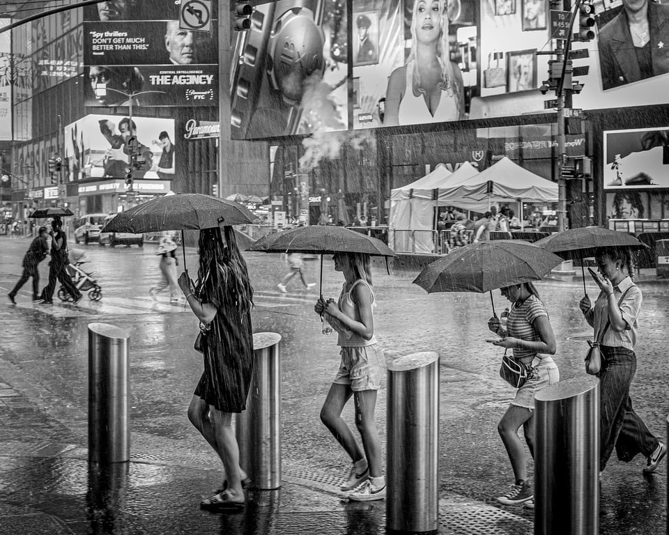 Pedestrians carrying umbrellas through a rainy city street, photographed in black and white.